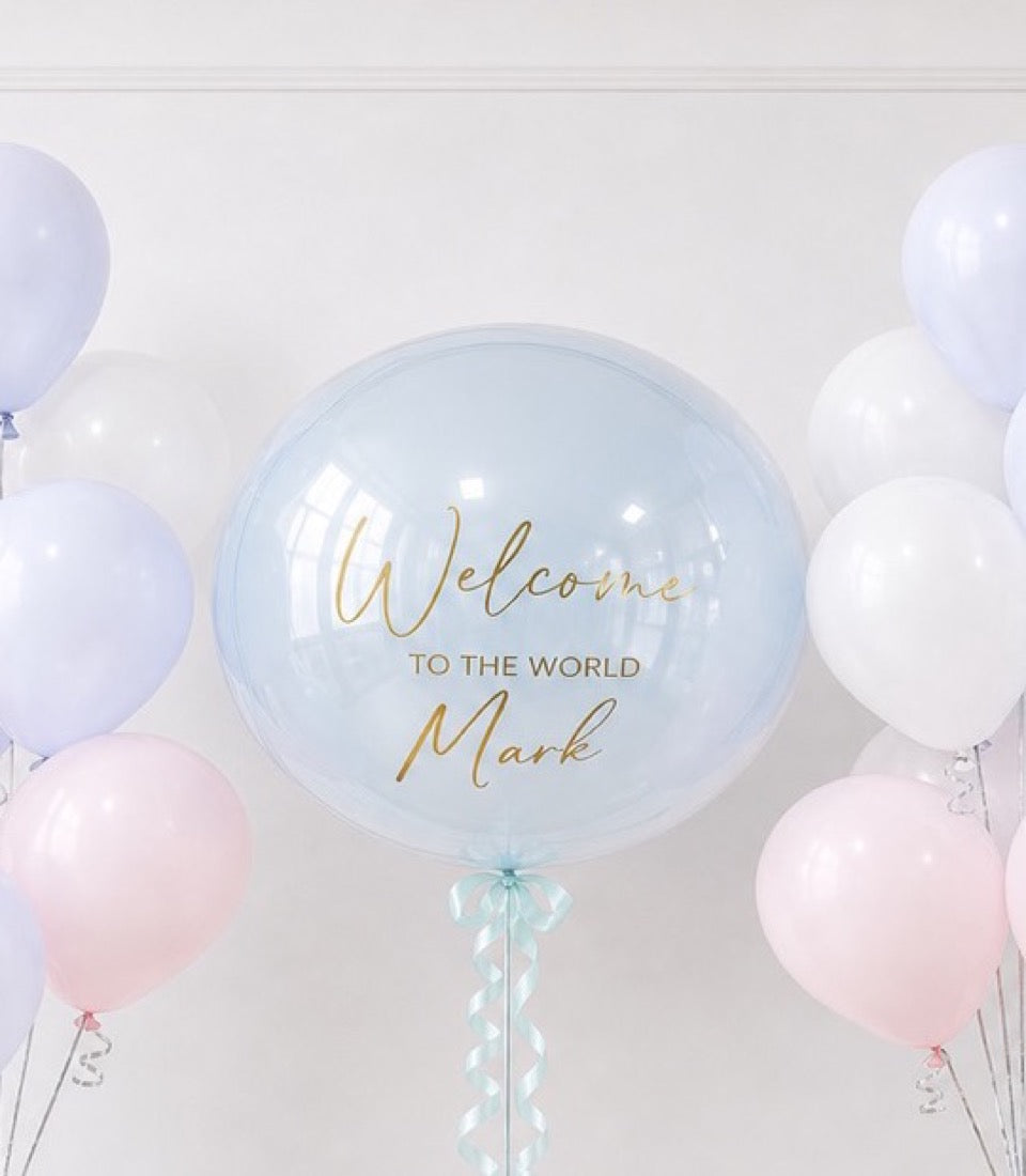 Large clear bubble balloon with gold "Welcome to the World Mark" text, surrounded by pastel pink, white, and lavender balloons against white ceiling.