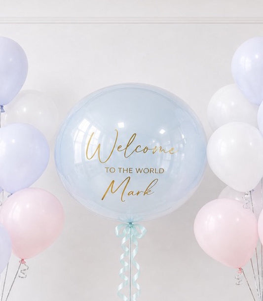 Large clear bubble balloon with gold "Welcome to the World Mark" text, surrounded by pastel pink, white, and lavender balloons against white ceiling.