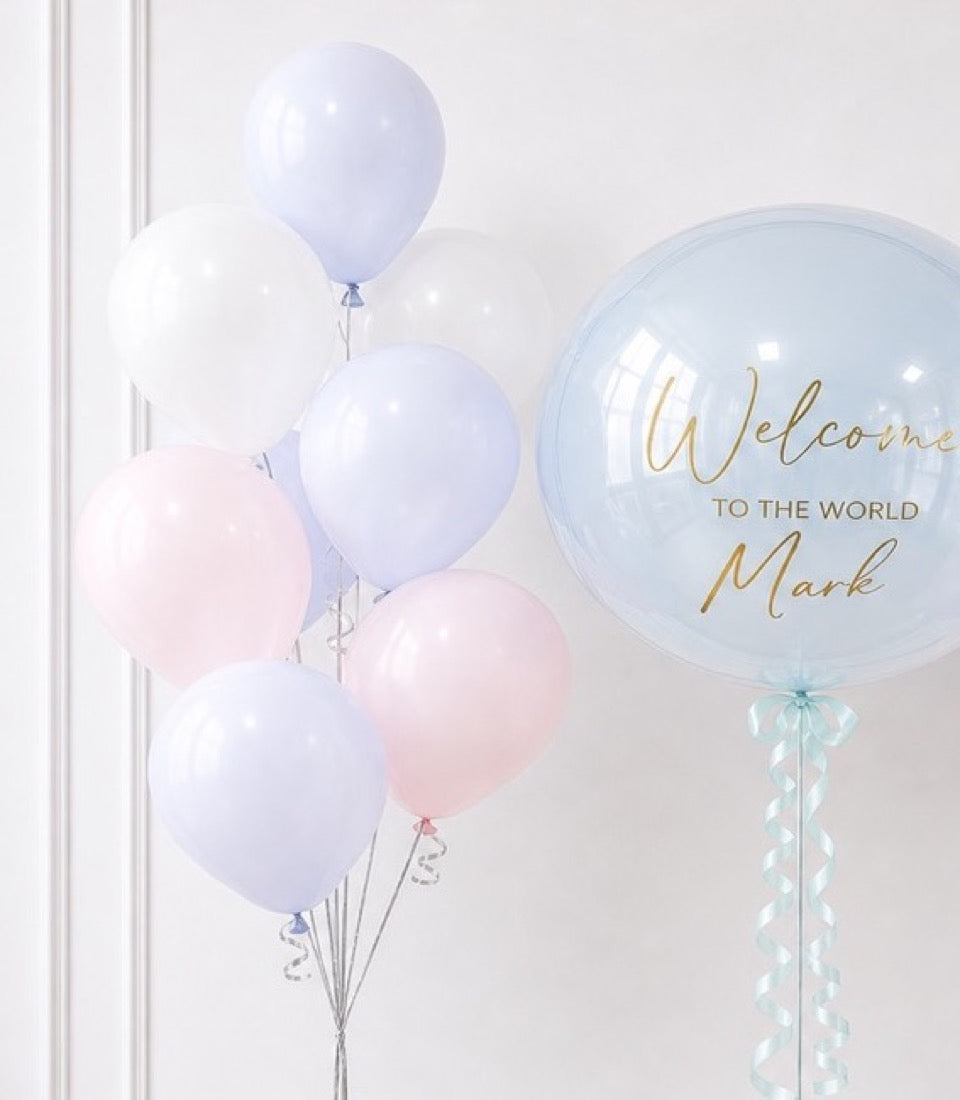 Pastel balloon arrangement with large bubble balloon reading "Welcome to the World Mark" in gold script, featuring lavender, white, and pink latex balloons with curled ribbons.