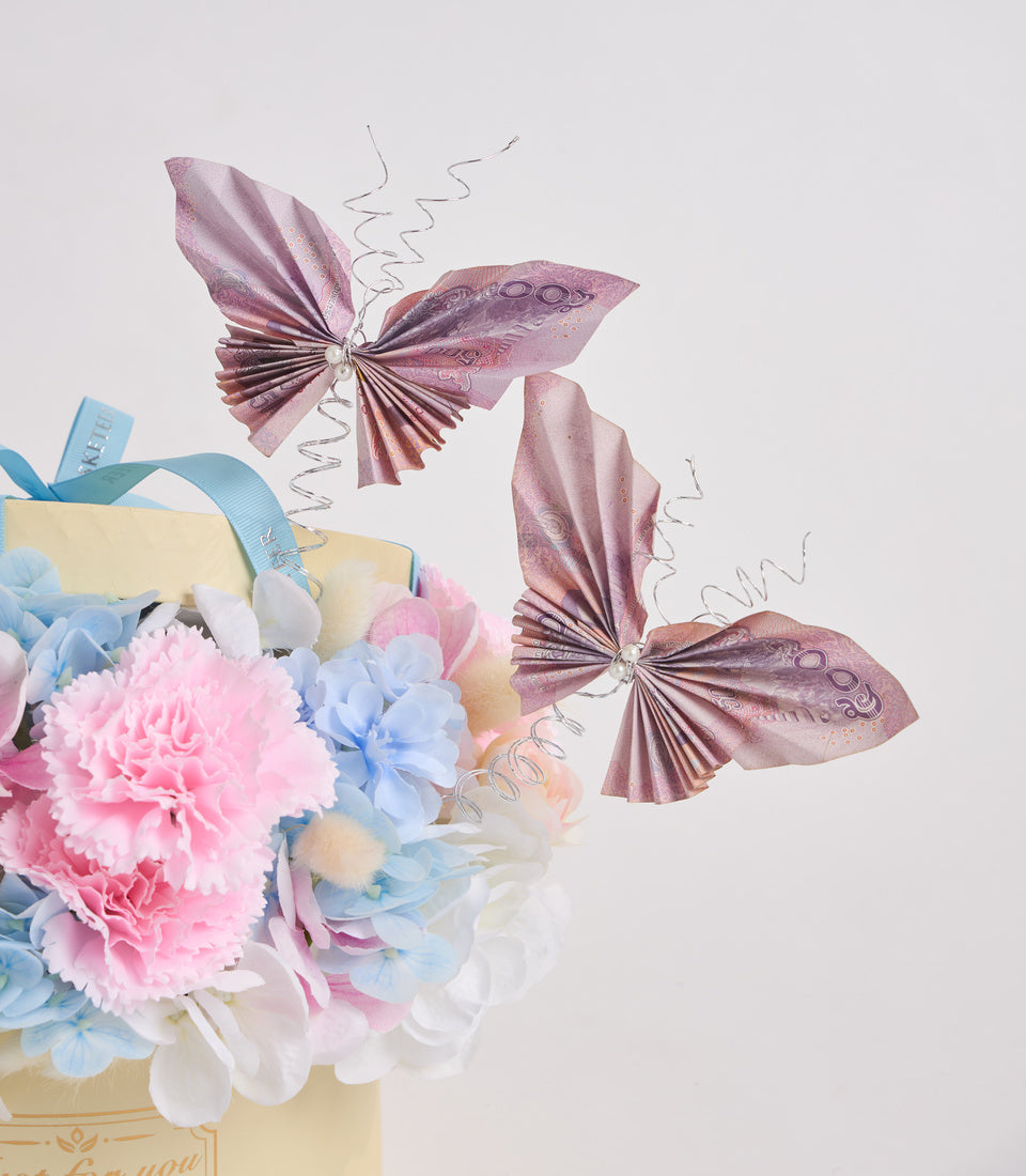 Elegant floral arrangement with pink carnations, blue hydrangeas, cream roses in a pale yellow box with blue ribbon, adorned with mauve decorative butterflies and silver wire accents.