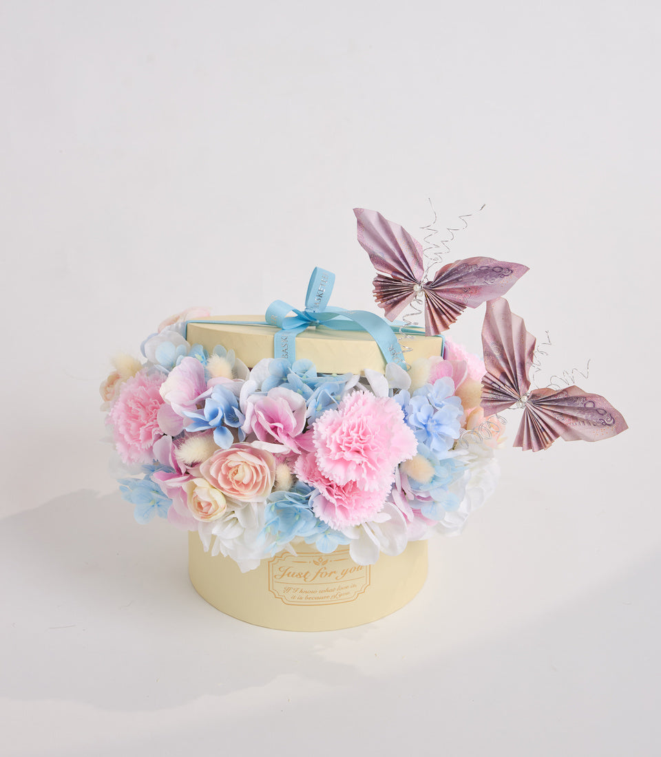 Luxury cream gift box adorned with pink carnations, blue hydrangeas, peach roses, decorative butterflies, and turquoise ribbon bow.