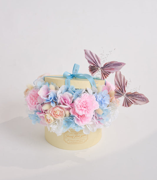 Luxury cream gift box adorned with pink carnations, blue hydrangeas, peach roses, decorative butterflies, and turquoise ribbon bow.