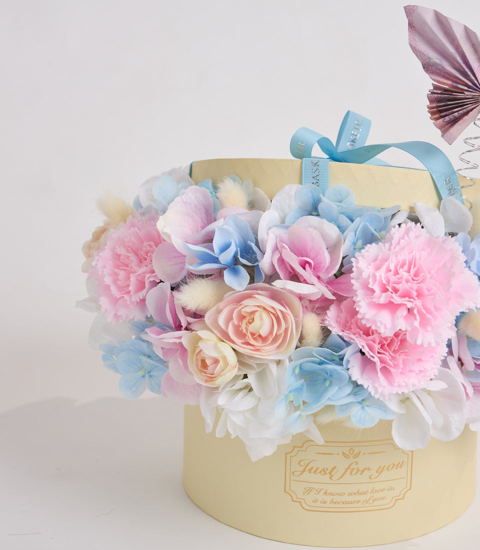 Luxury floral gift box with pink peonies, blue hydrangeas, cream roses, and white flowers in pale yellow cylindrical box with blue ribbon.