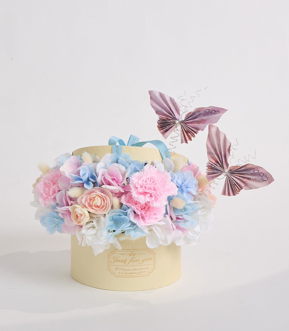 # Alt Text

Luxury cream cylindrical hatbox with "Just for you" label, filled with pink peonies, blue hydrangeas, peach roses, and white flowers, topped with decorative purple fabric butterflies.
