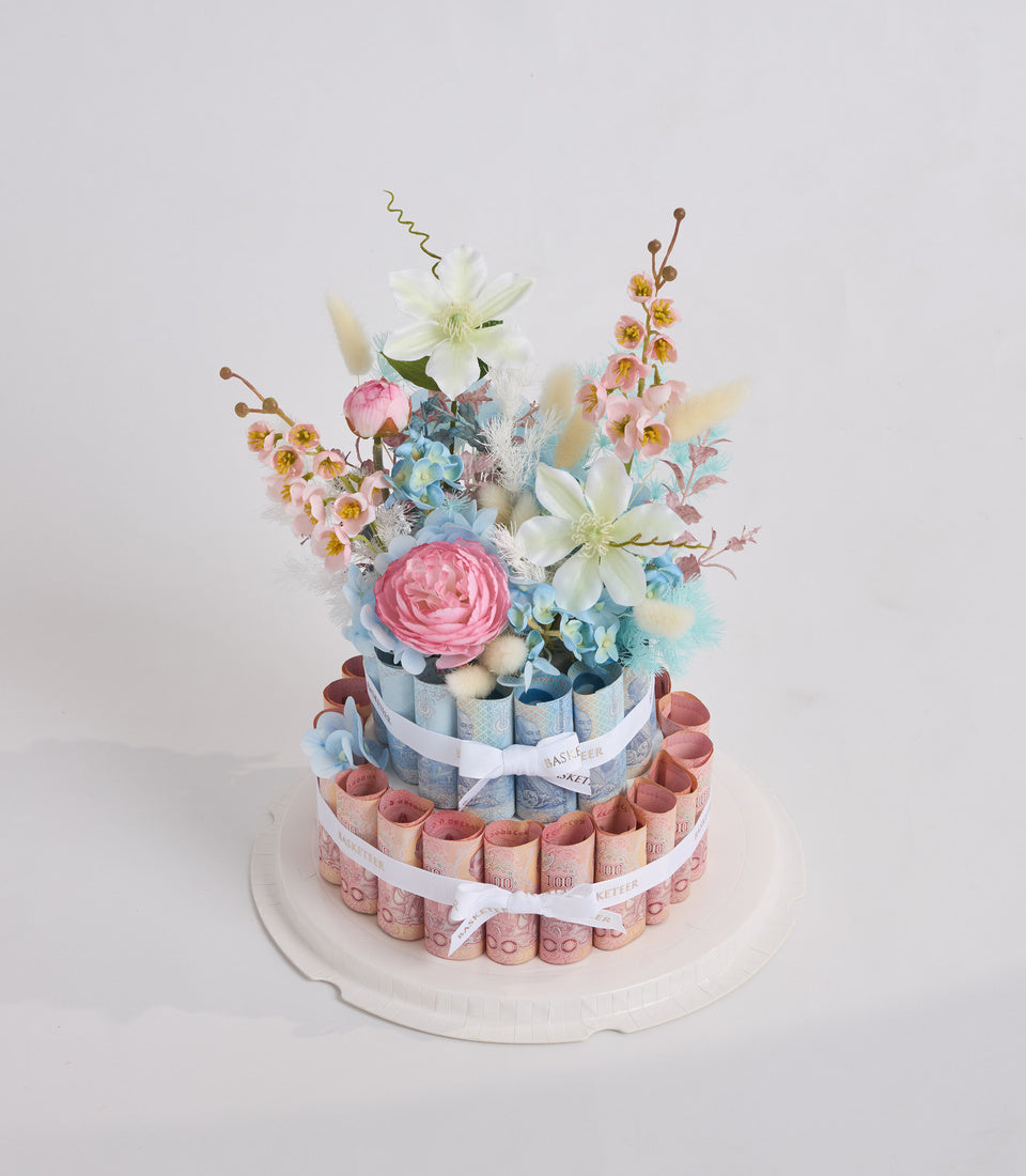 Two-tiered money cake with pink and blue rolled banknotes, topped with fresh flowers including pink roses, white clematis, and small yellow blooms on white plate.