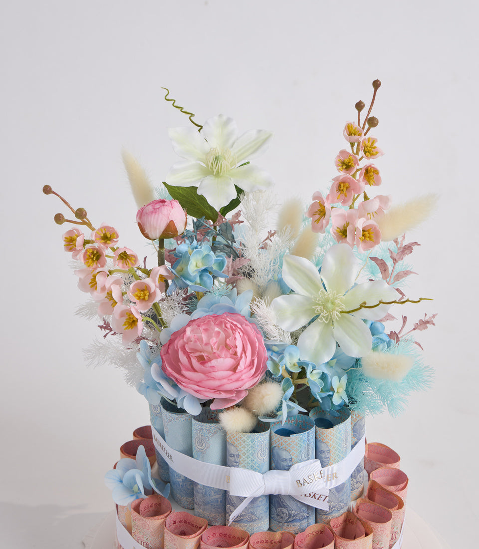 # Alt Text

Luxurious tiered flower arrangement with pink roses, white clematis, blue hydrangeas, and small pink flowers, wrapped in decorative blue and pink paper with white ribbon.