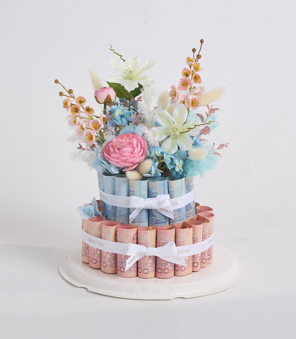 Two-tiered diaper cake with pink and blue rolled diapers, decorated with fresh flowers including pink roses, white clematis, blue hydrangeas, and small pink orchids, white ribbons.