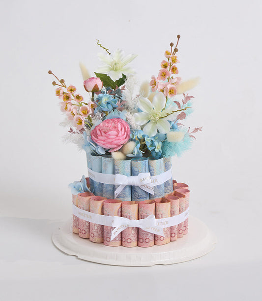 Two-tiered diaper cake with pink and blue rolled diapers, decorated with fresh flowers including pink roses, white clematis, blue hydrangeas, and small pink orchids, white ribbons.