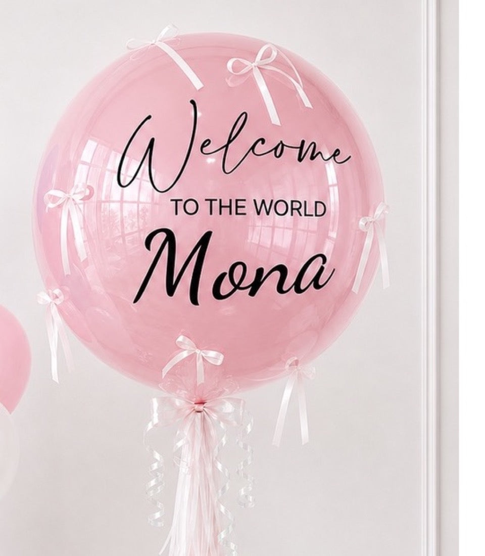 Pink bubble balloon with white ribbons and bows, printed text reading "Welcome to the World Mona" in black script and sans-serif fonts.