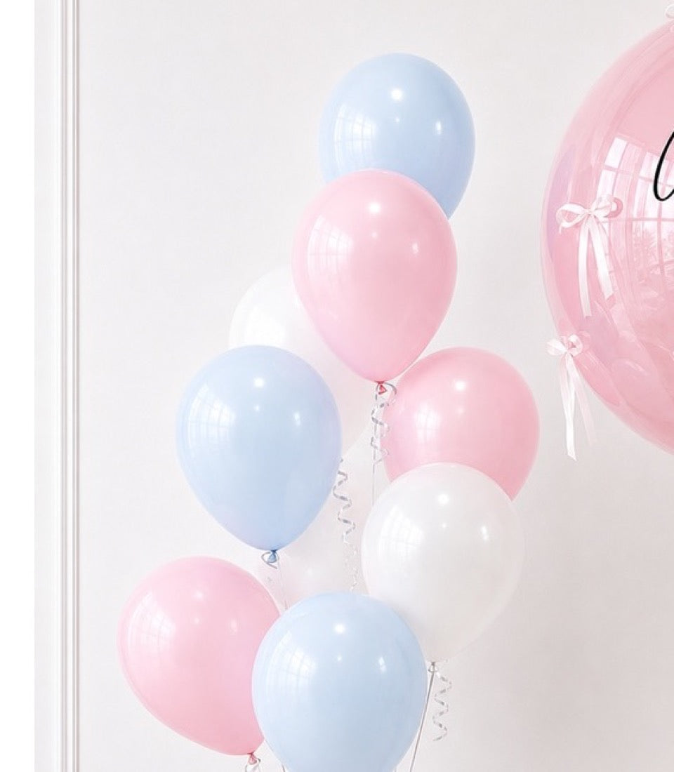 Pink, white, and light blue balloons with curling ribbons arranged vertically against a white wall, with a large pink tulle balloon visible on the right.