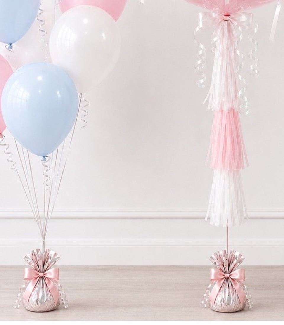 Pastel pink and blue balloons with white tulle tassels and pink ribbon bows on wooden floor against white wall.