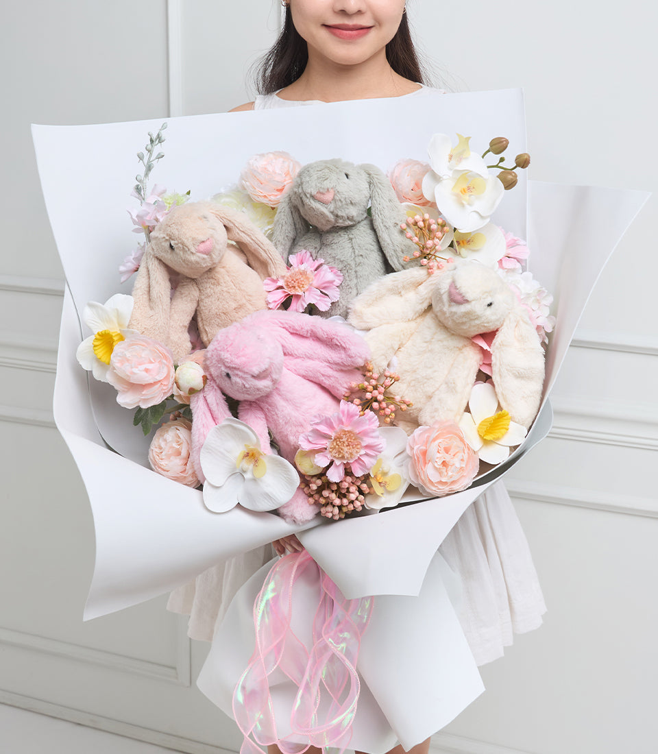 Luxurious plush bunny bouquet with soft gray, pink, and cream rabbits nestled among fresh roses, peonies, and white flowers with gold accents, wrapped in white paper with pink ribbon.