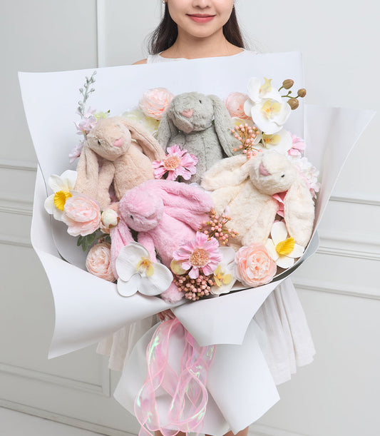 Luxurious plush bunny bouquet with soft gray, pink, and cream rabbits nestled among fresh roses, peonies, and white flowers with gold accents, wrapped in white paper with pink ribbon.