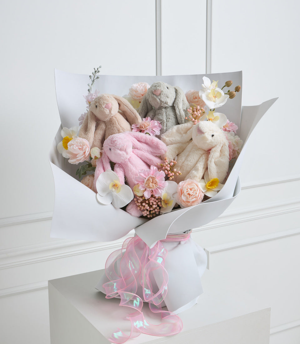 Luxury gift bouquet featuring three plush stuffed bunnies in pink, gray, and cream surrounded by fresh flowers including roses, orchids, and peonies, wrapped in white paper with pink ribbon.