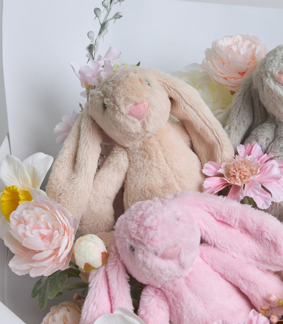 Soft plush bunny rabbits in cream and pink surrounded by delicate flowers including peonies, roses, and daisies.
