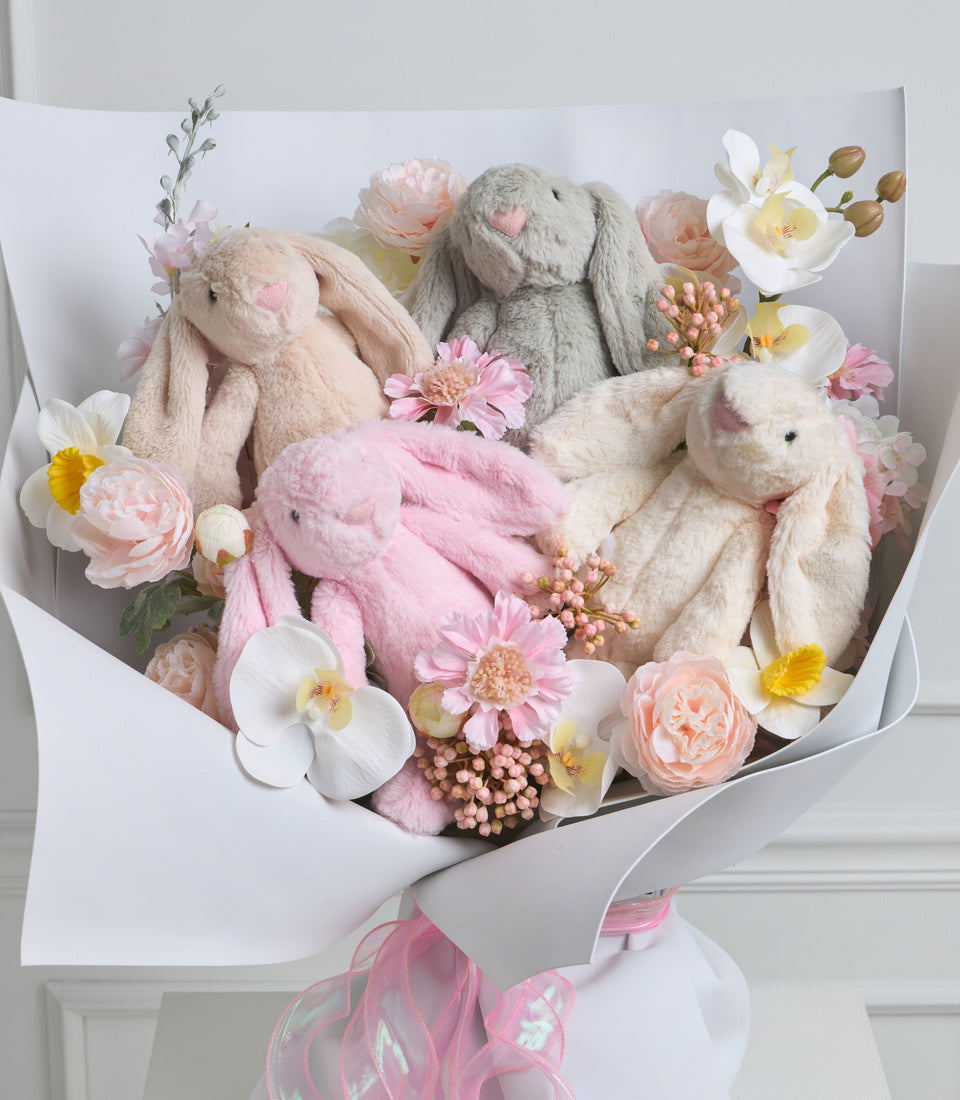 Luxe gift bouquet featuring four plush stuffed bunnies in cream, pink, and gray nestled among fresh roses, peonies, daisies, and greenery, wrapped in white paper with pink ribbon.