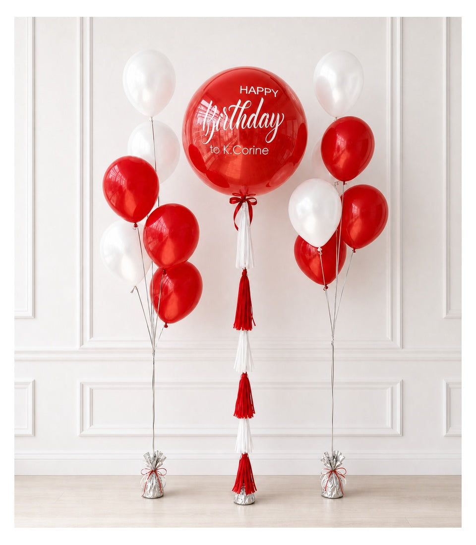 Red and white birthday balloon bouquet with large red orbz balloon reading "Happy Birthday to K.Corine" and tassel decorations on white paneled wall.