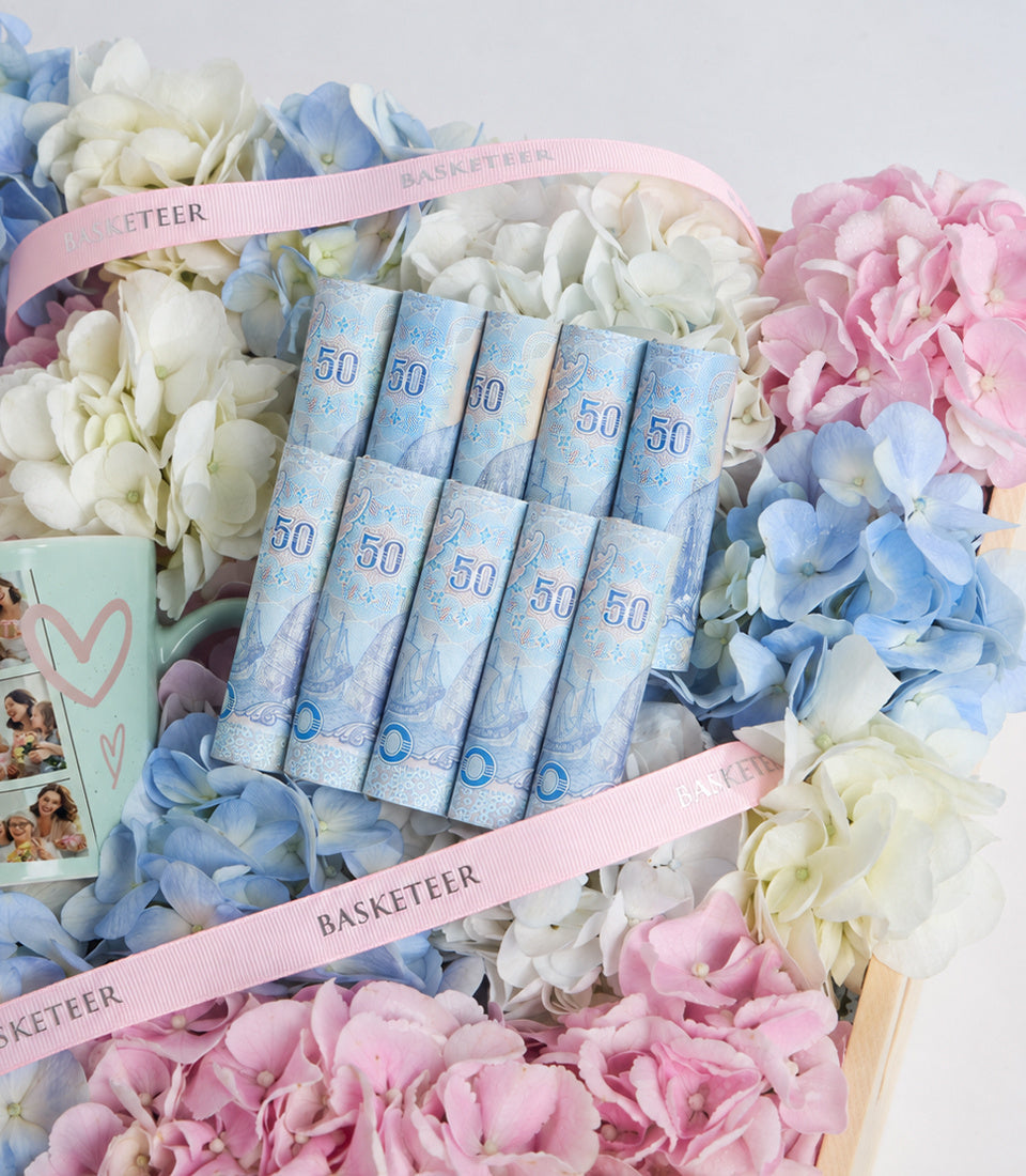 # Alt Text

Blue £50 note napkins arranged in a gift box with pink ribbons, surrounded by white, pink, and blue hydrangea flowers.