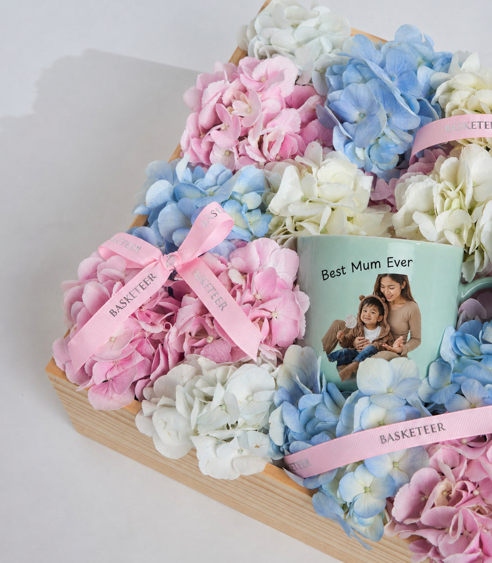 # Alt Text

Personalized "Best Mum Ever" mug surrounded by pink, blue, and white hydrangeas in a wooden basket with pink ribbons.