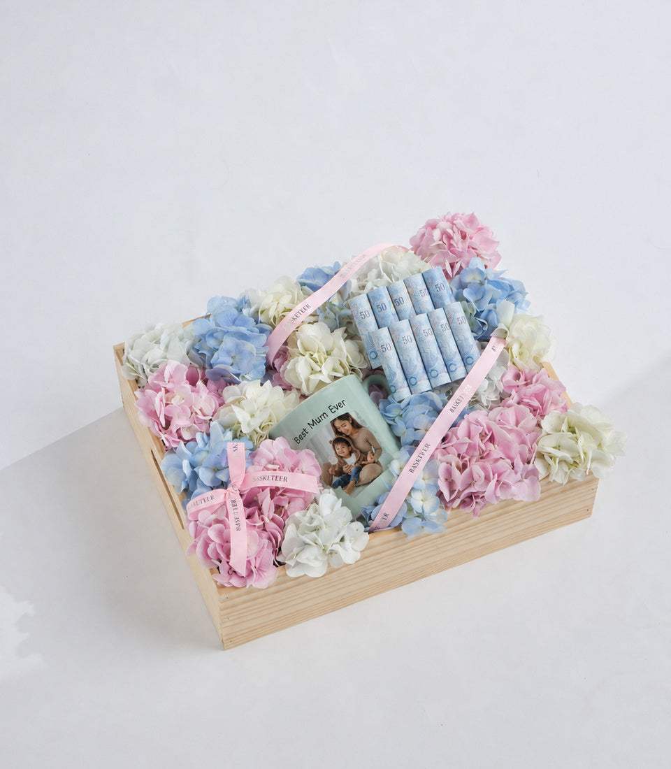 Wooden box arrangement with pink, blue, and white hydrangeas, blue skincare bottles with pink ribbons, and a "Best Mum Ever" card on white background.