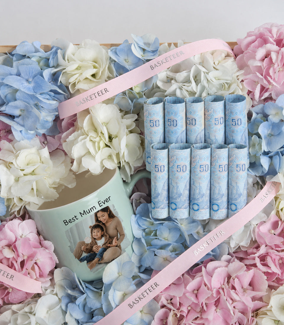 Luxury gift set featuring blue patterned tissues marked "50", a personalized "Best Mum Ever" mug, and pink ribbon, surrounded by pink, blue, and white flowers.
