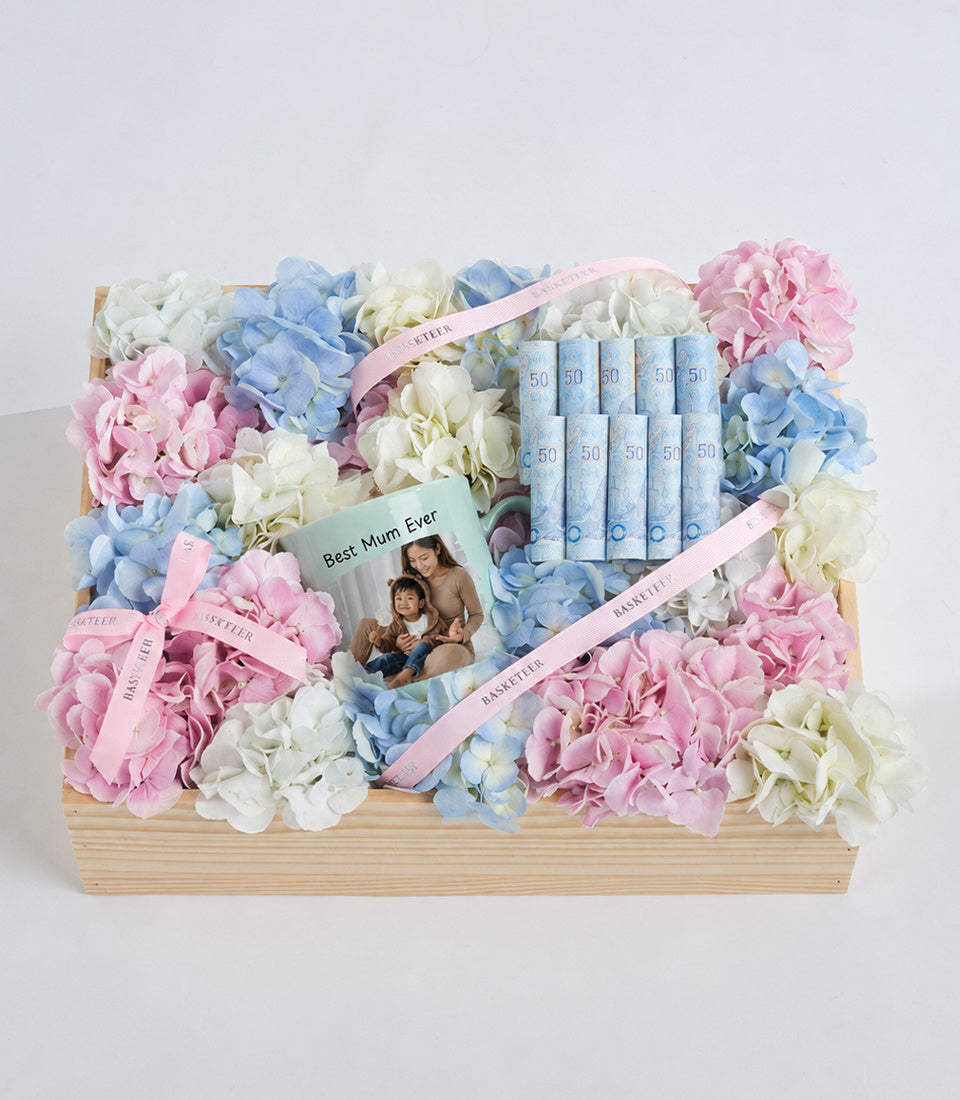 Luxury gift basket with pink, blue, and white hydrangeas in wooden box, containing skincare products, SPF 50 sunscreen tubes, photo frame card labeled "Best Mum Ever" and pink ribbons.