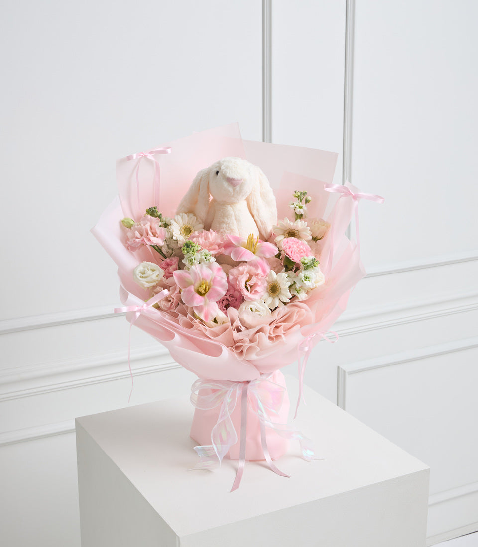 Pink flower bouquet with cream bunny plush toy, wrapped in pink paper with ribbon bows, displayed on white pedestal.