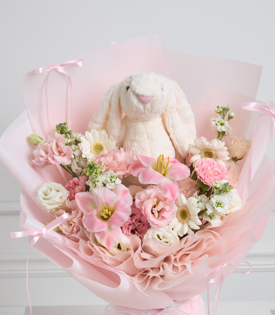 Pink wrapped bouquet with white plush bunny, pink and white flowers including daisies and carnations, tied with pink ribbons.