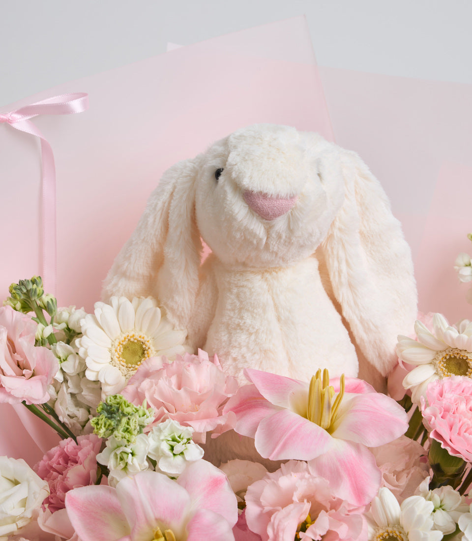 Cream plush bunny toy surrounded by pink and white flowers against a pink background with ribbon detail.