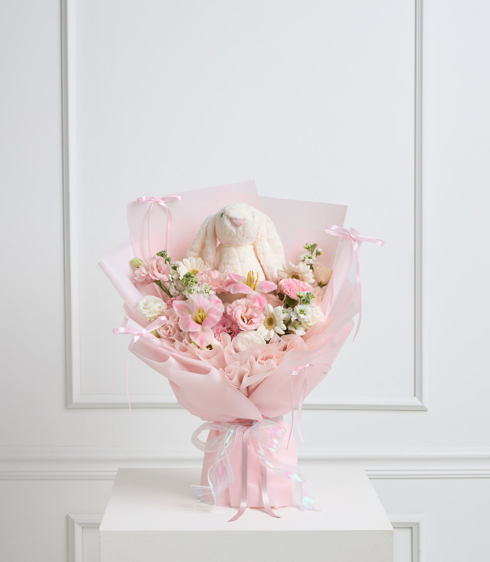 Elegant pink bouquet with cream bunny plush, pink and white flowers, wrapped in pink paper with ribbons, displayed on white pedestal.