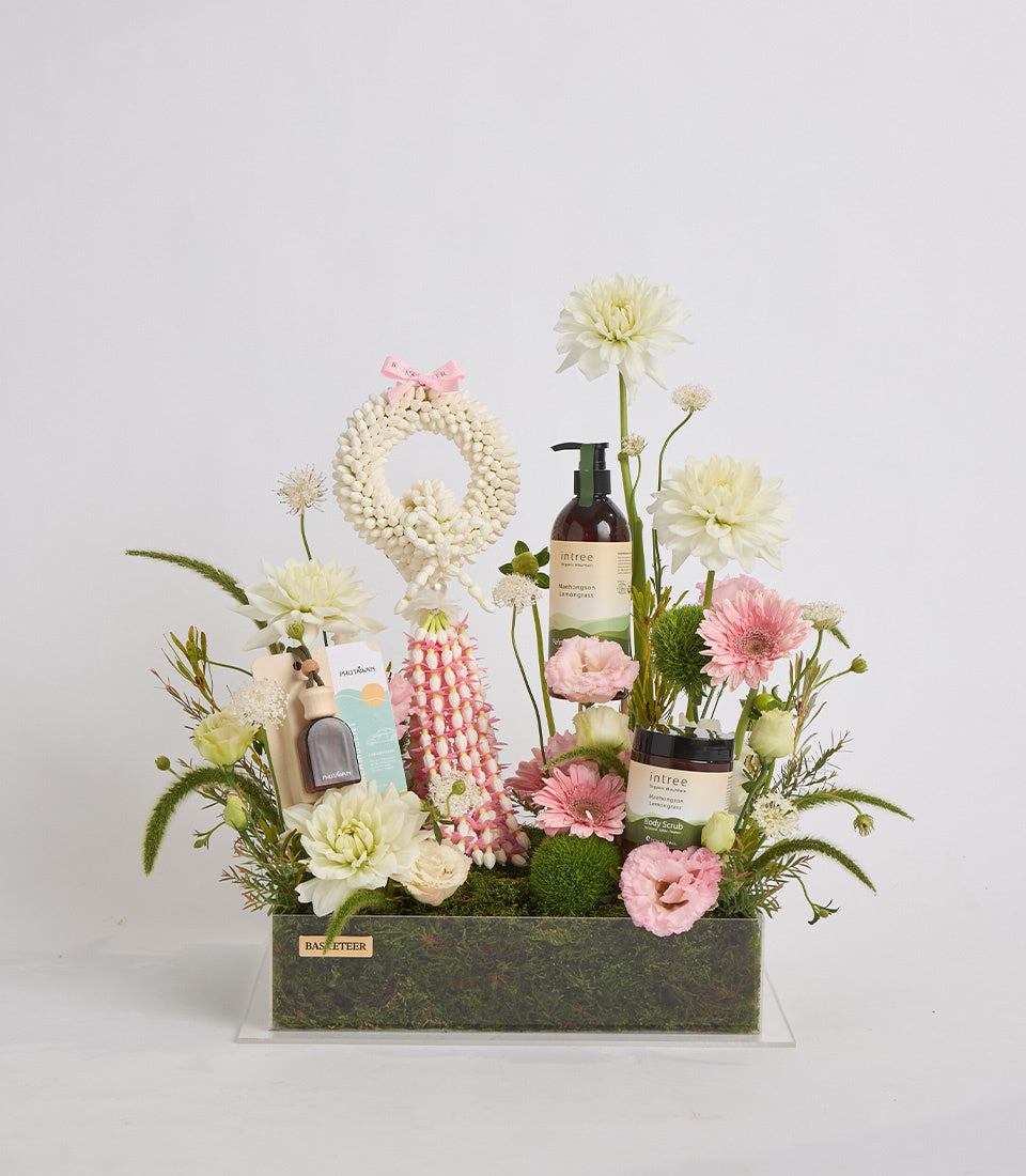 Luxe spa gift arrangement featuring cream and pink flowers, beaded wreath with pink bow, Intree skincare bottles, body scrub jar, and decorative accessories in textured green container.
