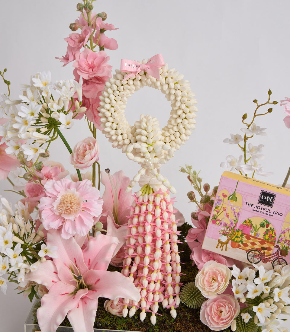 Floral arrangement with cream pearl beaded wreath, pink and white flowers, gift box, and pink bow on gray background.
