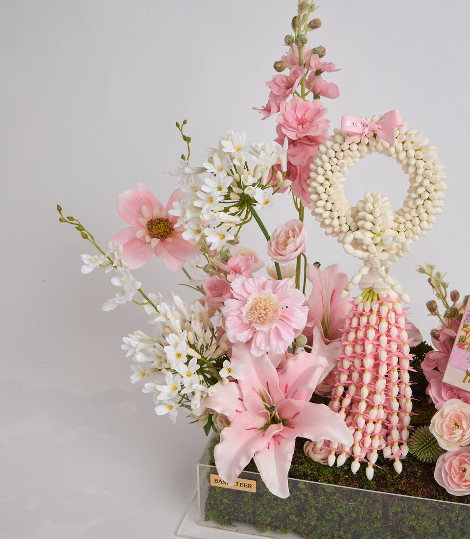 Luxurious floral arrangement in pink and white with pearl beaded wreaths, daisies, roses, and lilies in a moss-lined container.
