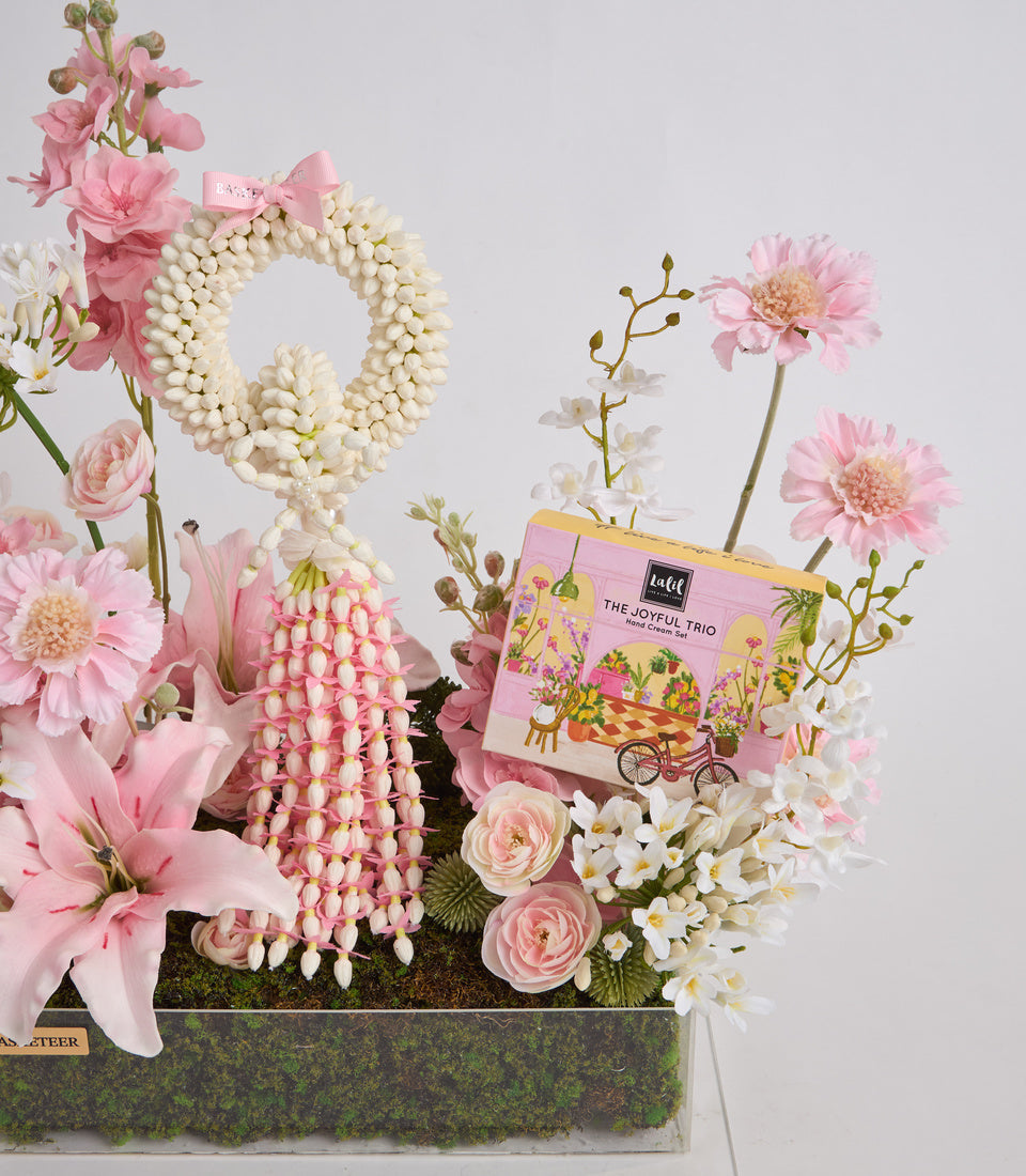 Luxe floral gift arrangement with pink gerberas, white pearls, cream flowers, and "The Joyful Trio" product box in modern botanical display.