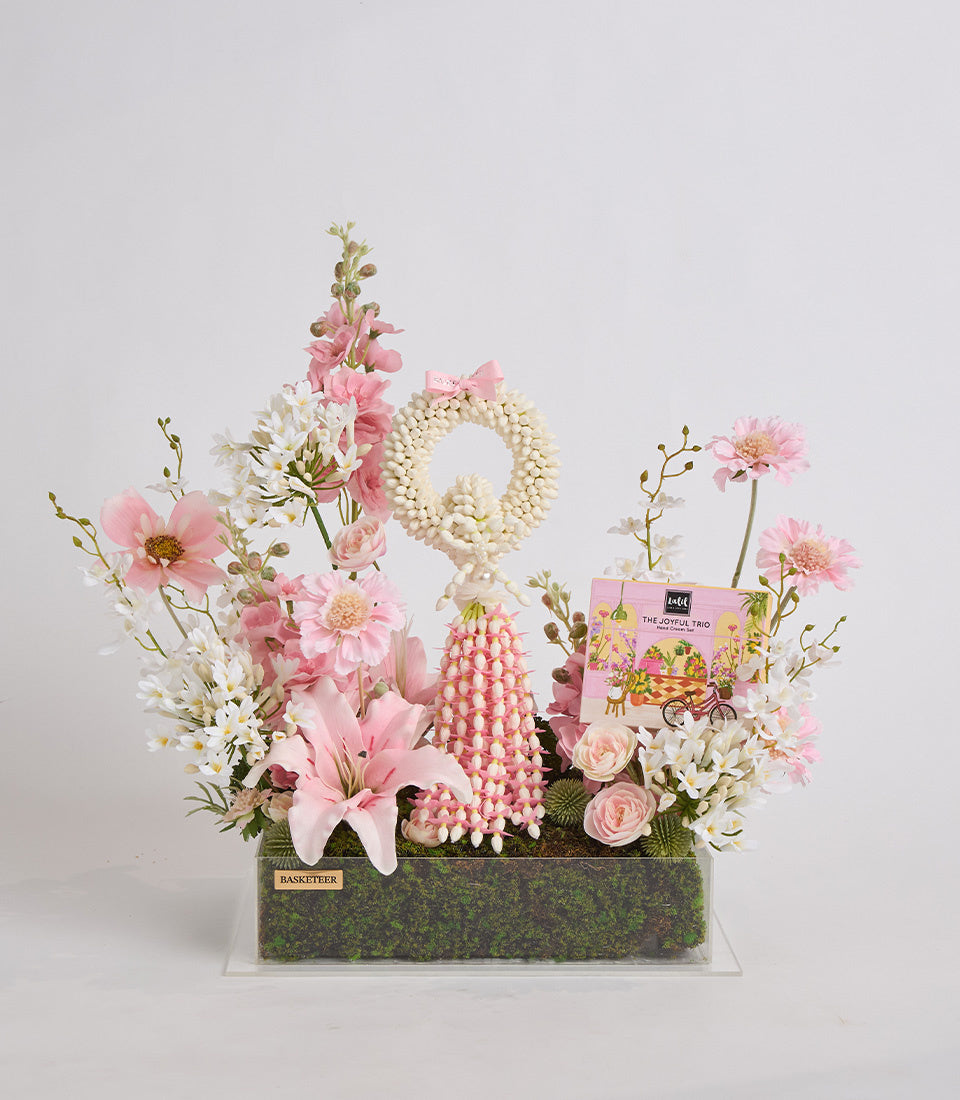 Luxurious floral arrangement featuring pink and white flowers, cream pearl beads, and decorative elements in a moss-lined green container with gift card.