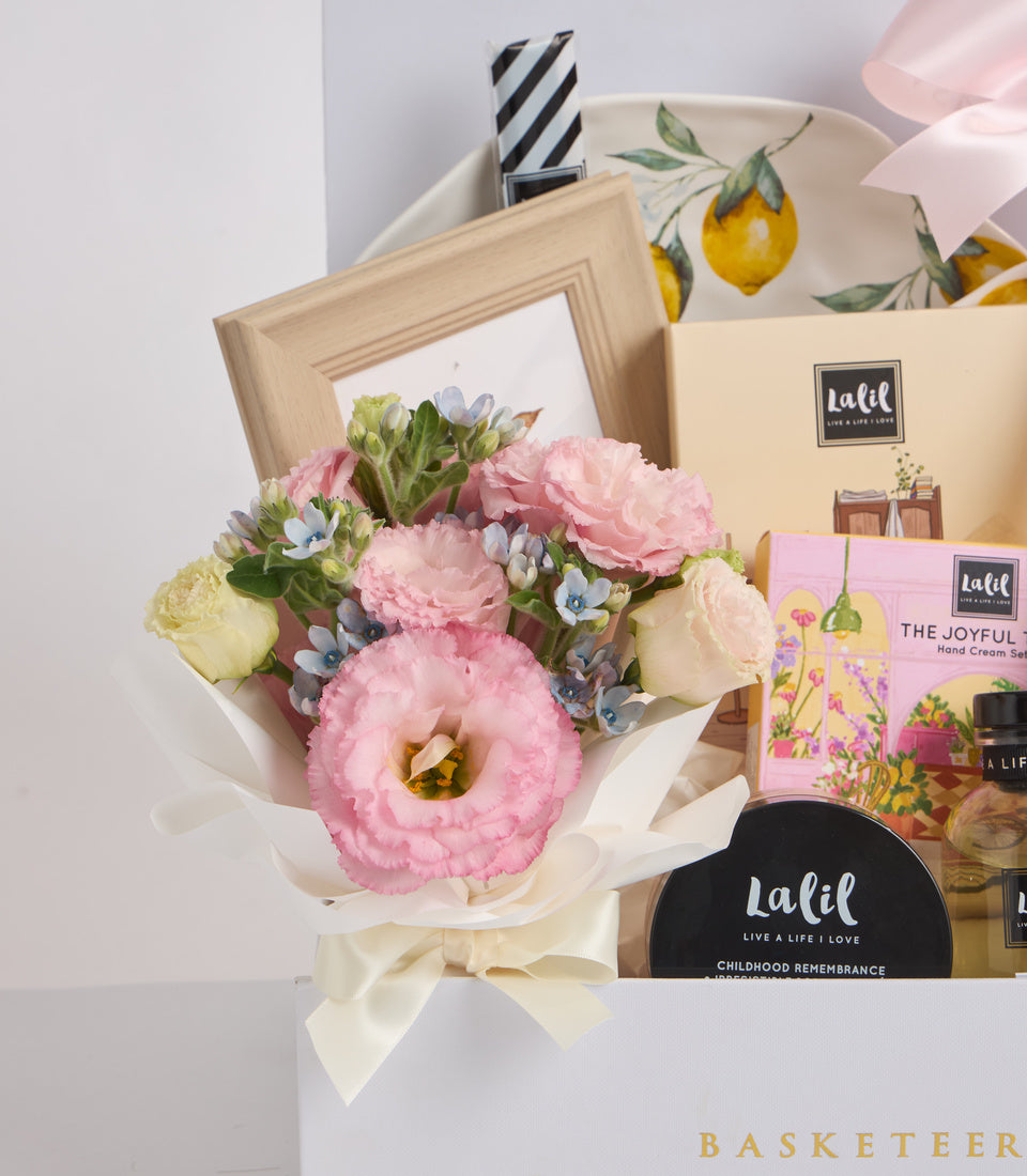 Luxury gift basket with pink and cream flowers, lemon-patterned ceramic bowl, Lalil beauty products, wooden frame, and striped candle on white background.