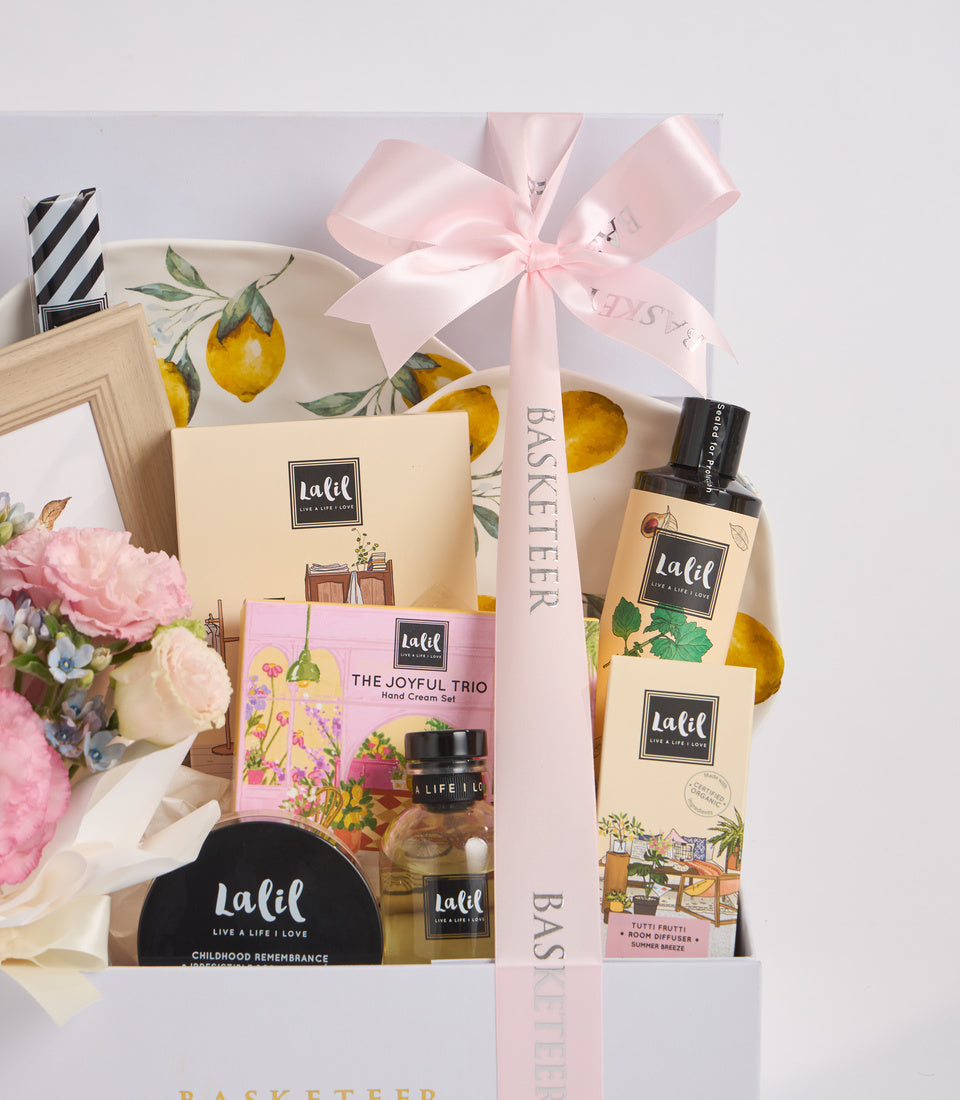 Elegant gift basket featuring Lalil beauty products, lemon-patterned plate, pink flowers, wooden frame, and pink ribbon bow on white background.