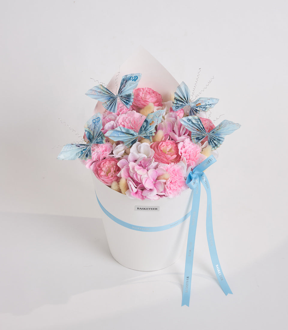 Luxurious floral arrangement in white cylindrical box with pink peonies, cream roses, blue decorative butterflies, and light blue ribbon bow.