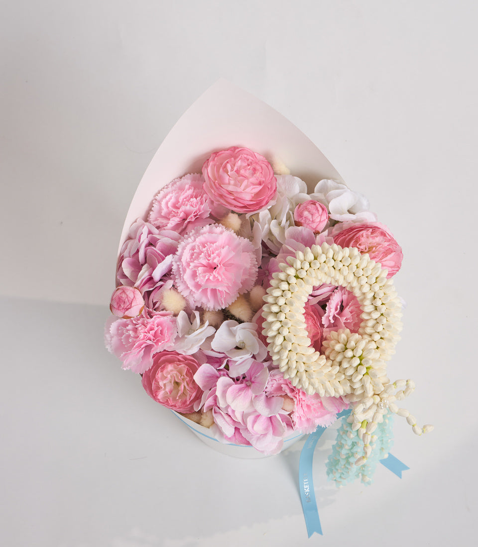 Luxury floral egg-shaped arrangement with pink peonies, white flowers, cream beaded strands, and blue ribbon bow on white background.