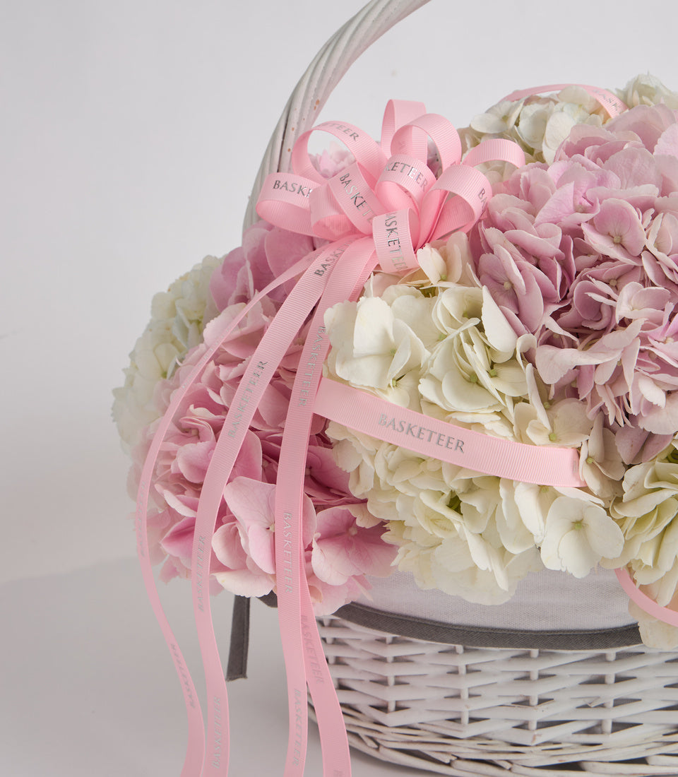 Elegant flower basket with pink and white hydrangeas, white wicker base, pink "Basketeer" ribbon bow, and gray fabric liner against neutral background.