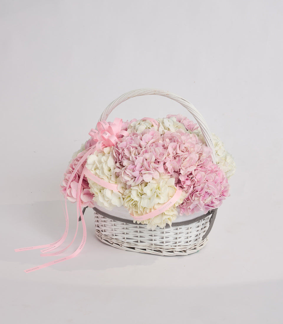 White wicker basket filled with pink and cream hydrangeas, tied with pink ribbon bow and clear handle, on white background.