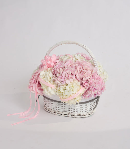 White wicker basket filled with pink and cream hydrangeas, tied with pink ribbon bow and clear handle, on white background.