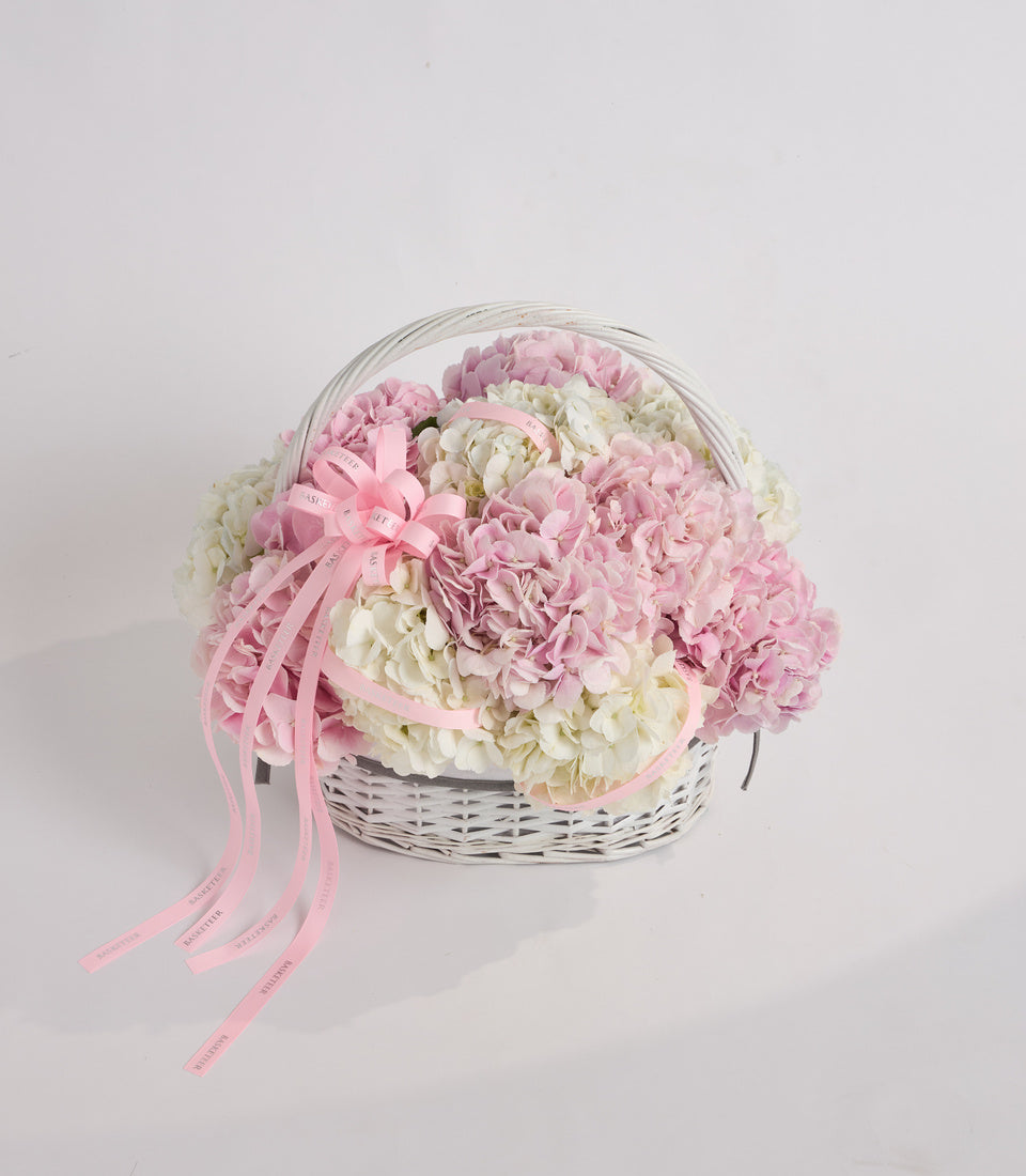 White wicker basket filled with pink and cream carnations, topped with a clear dome lid and pink ribbon bow.