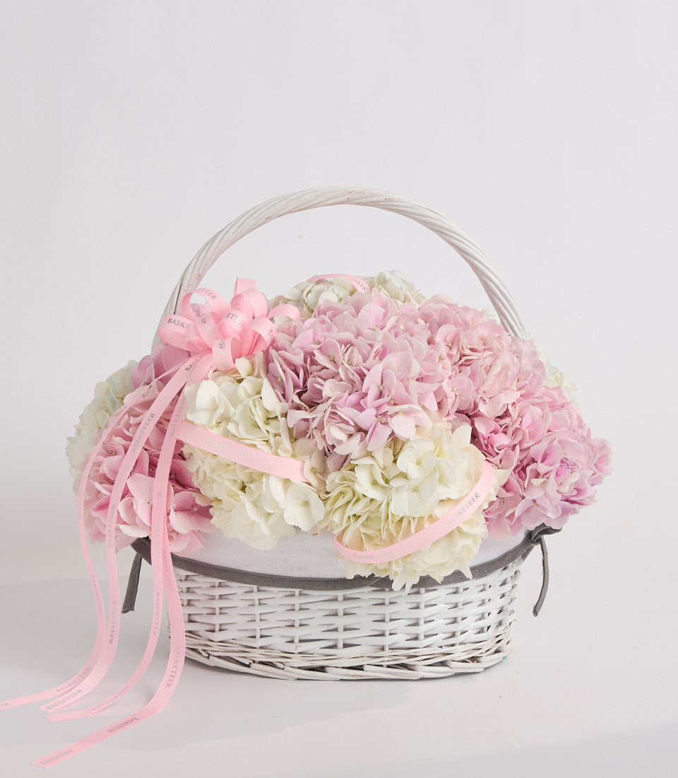 White wicker basket filled with pink and cream hydrangeas, decorated with pink ribbons and gray fabric liner.