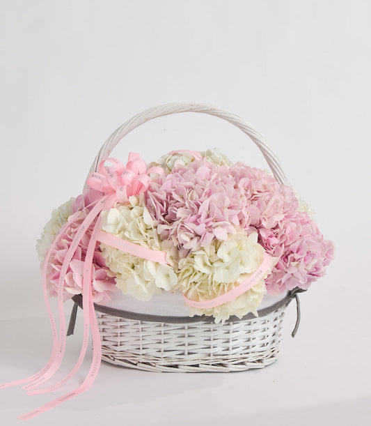 White wicker basket filled with pink and cream hydrangeas, decorated with pink ribbons and gray fabric liner.