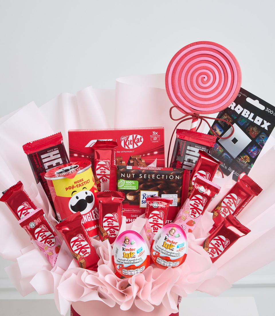 Red and pink candy gift basket with KitKat bars, Kinder eggs, lollipop, Roblox gift card, and pink tissue paper on white background.