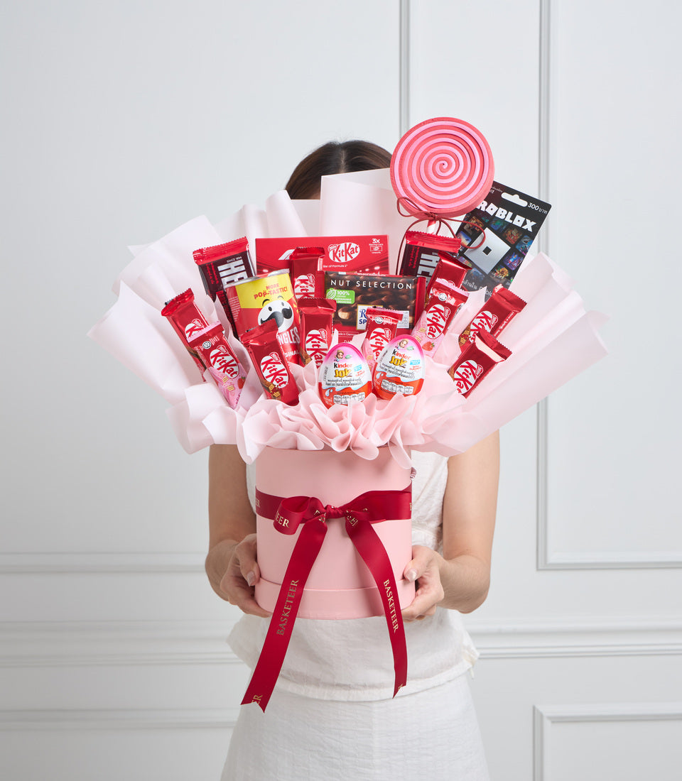 # Alt Text

Person holding large pink gift arrangement with KitKat candies, red ribbons, pink tissue paper, lollipop topper, and Roblox game card against white backdrop.