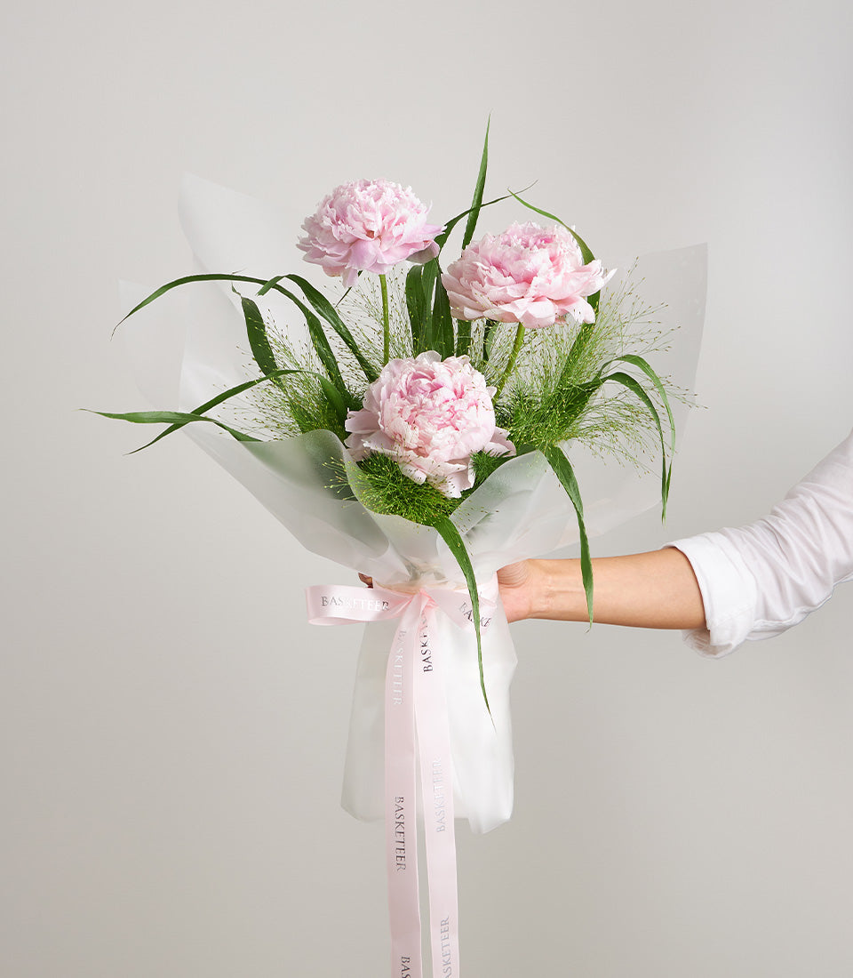 Elegant bouquet of three premium pink peonies with lush green foliage, wrapped in delicate
