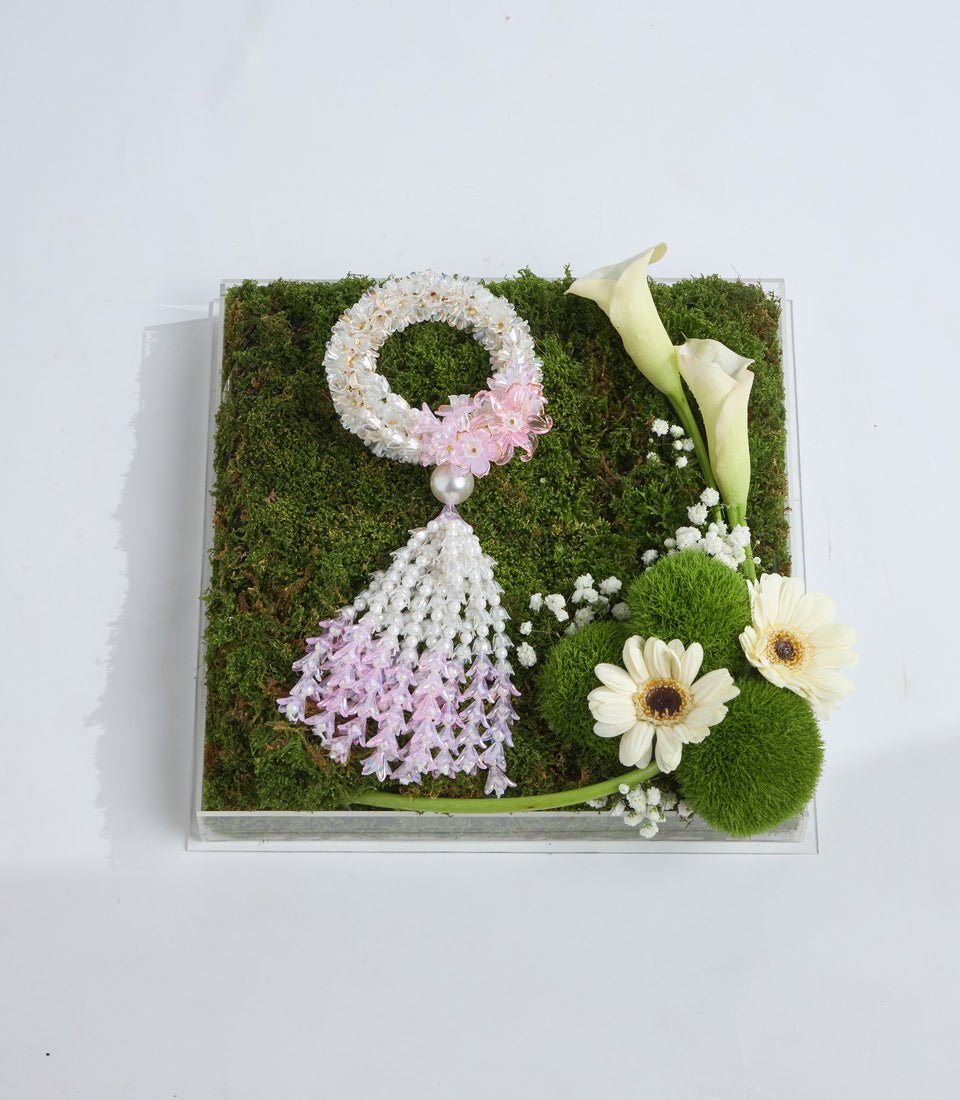 Elegant floral arrangement on moss base featuring white and pink flowers, calla lilies, gerbera daisies, baby's breath, and pearl accents in clear acrylic box.