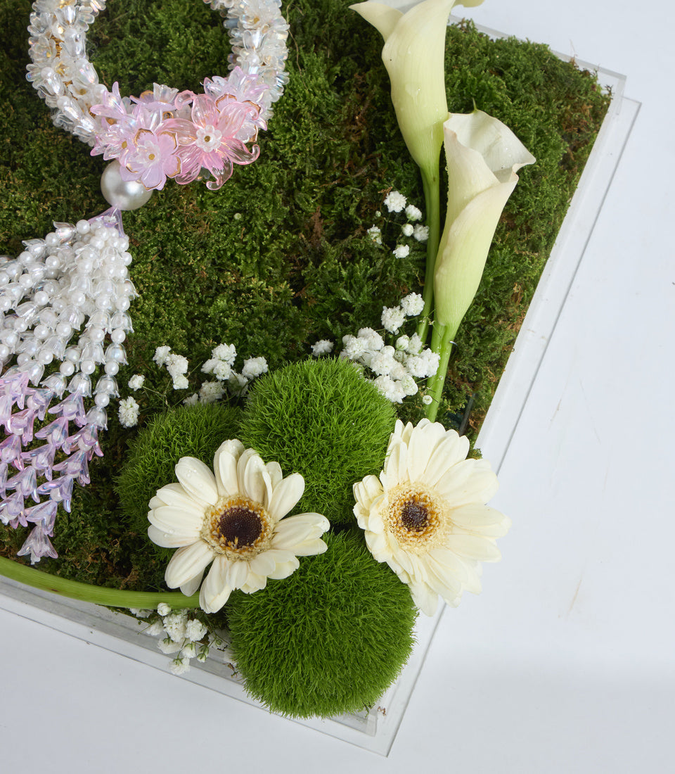 Floral arrangement in white frame featuring cream gerberas, pale yellow calla lilies, pink daisies, white baby's breath, moss base, and pearl accents.
